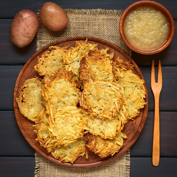 Golden-brown potato pancakes on a wooden plate, with applesauce, raw potatoes, and a fork.